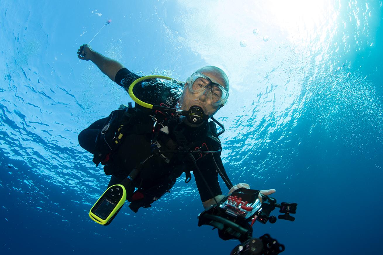A diver holding photography equipment stares into camera in shallow water.